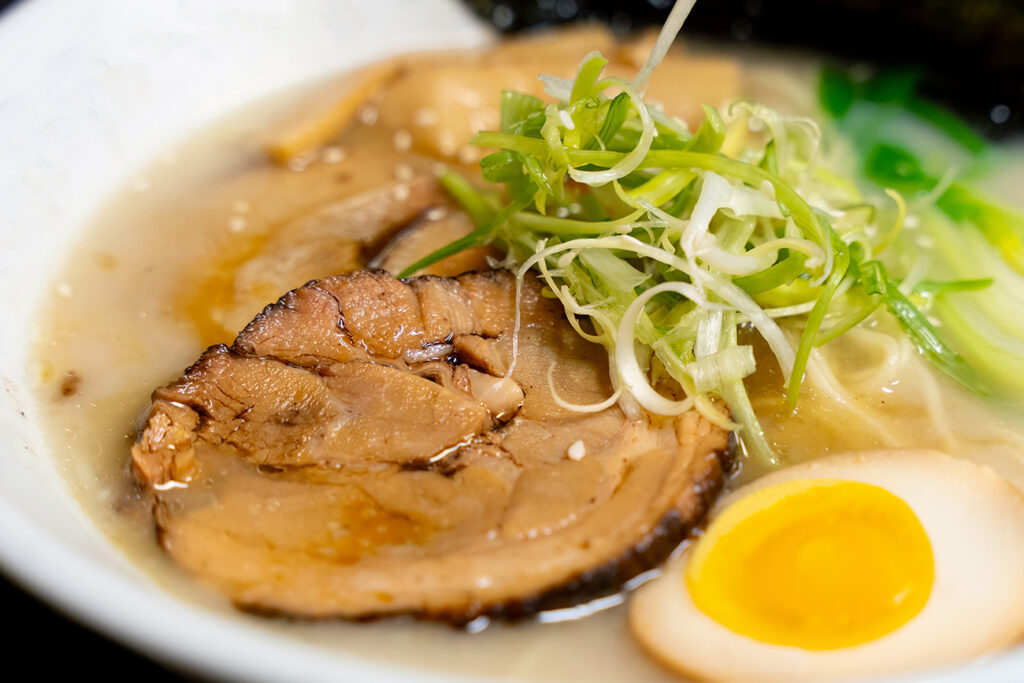 This image showcases a Tonkotsu ramen bowl of traditional Japanese ramen with slices of pork, halved boiled egg, seaweed, green onions, and other toppings, served in a white bowl.